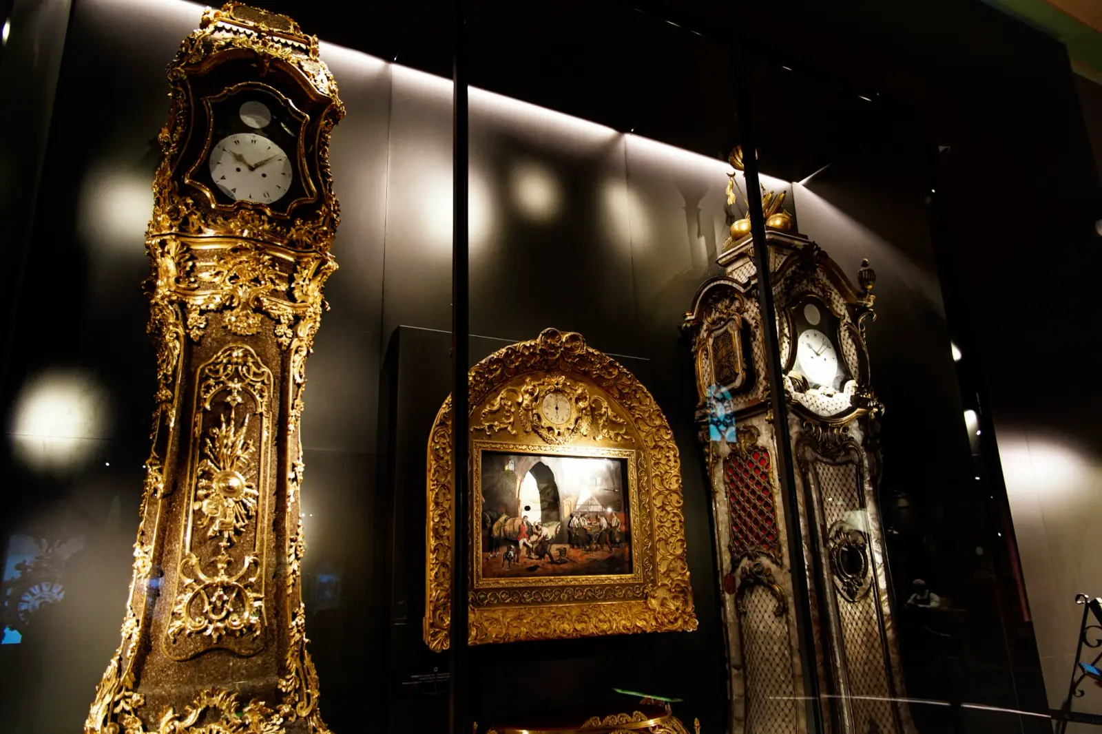 A gilded longcase clock in the Clock Museum, beside a painted panel.