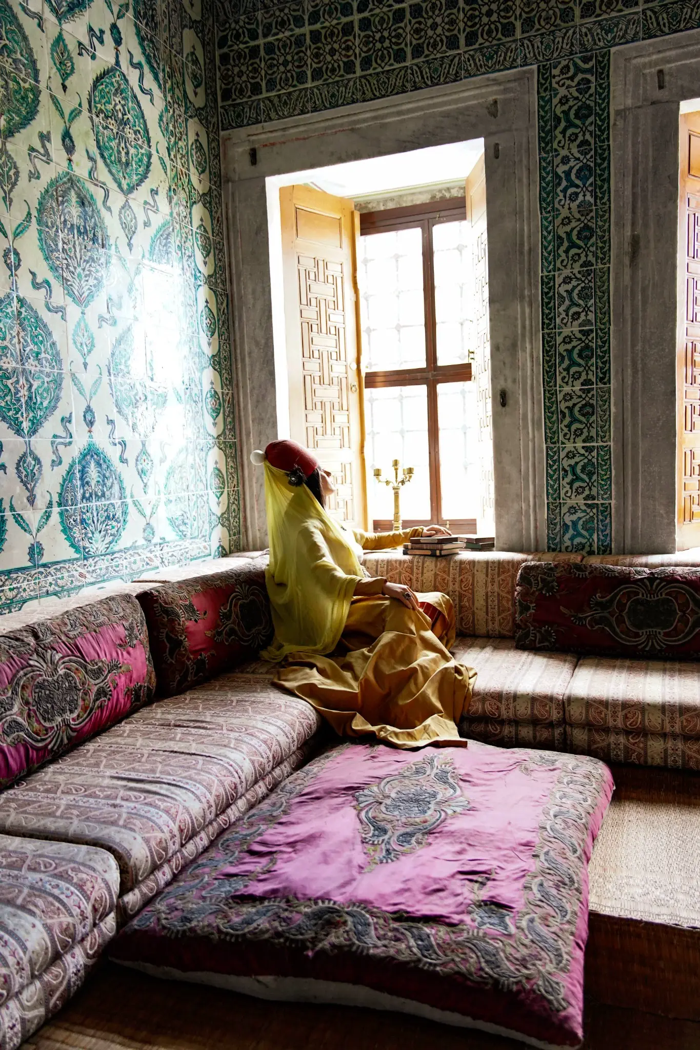 A costumed figure seated at a Harem window, İznik tiles and cushions around her.