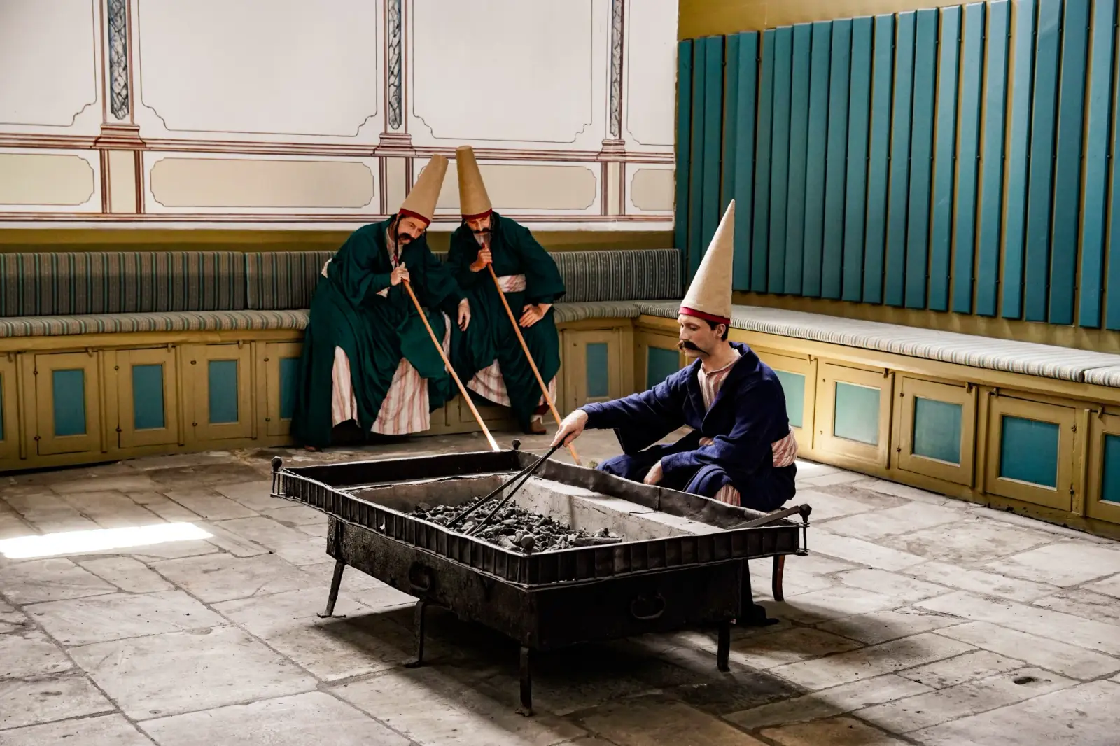 The Helvahane: costumed confectioners working a tray of macun with long paddles in the painted hall.